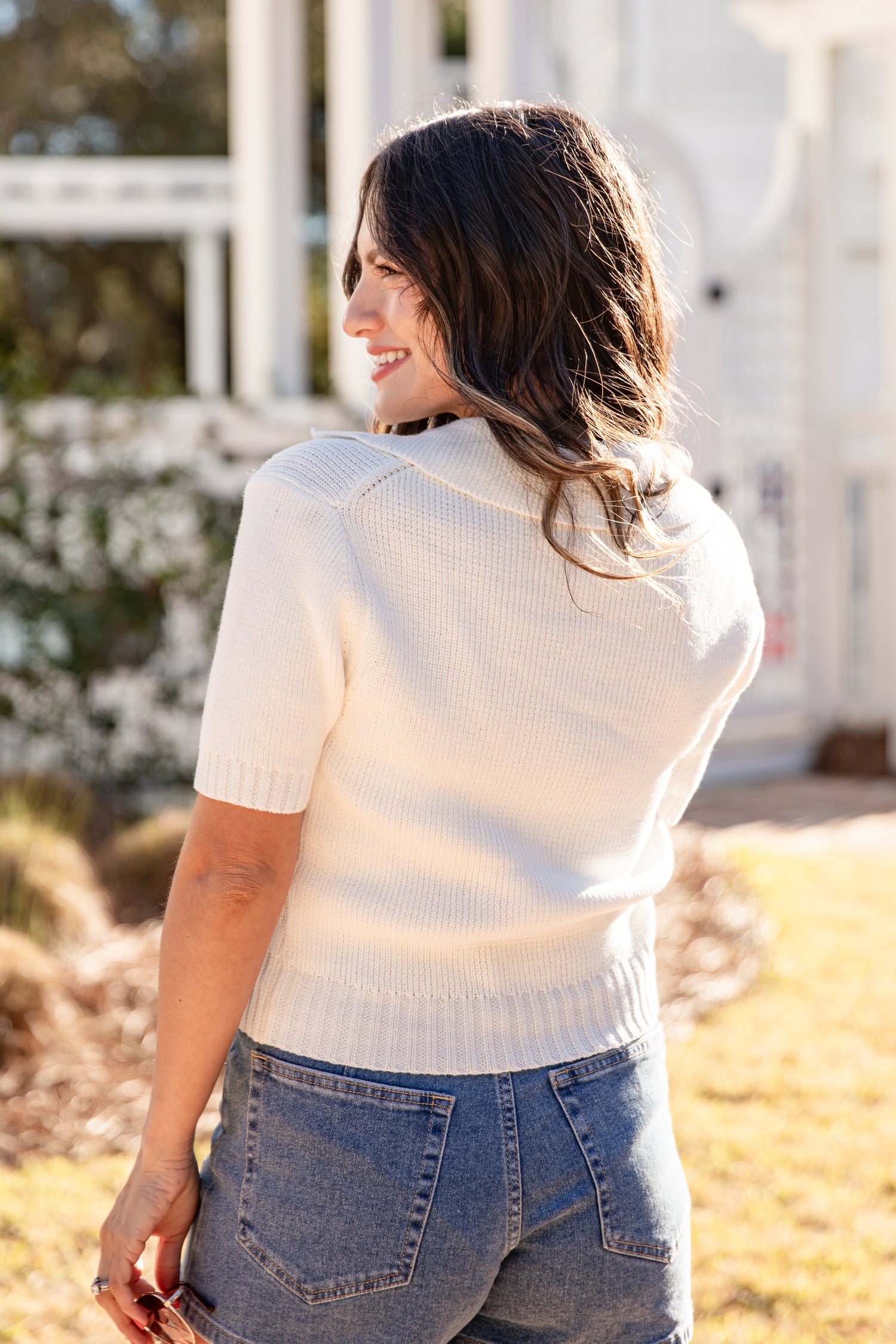 Woman wearing a white sweater and blue jeans standing outdoors.