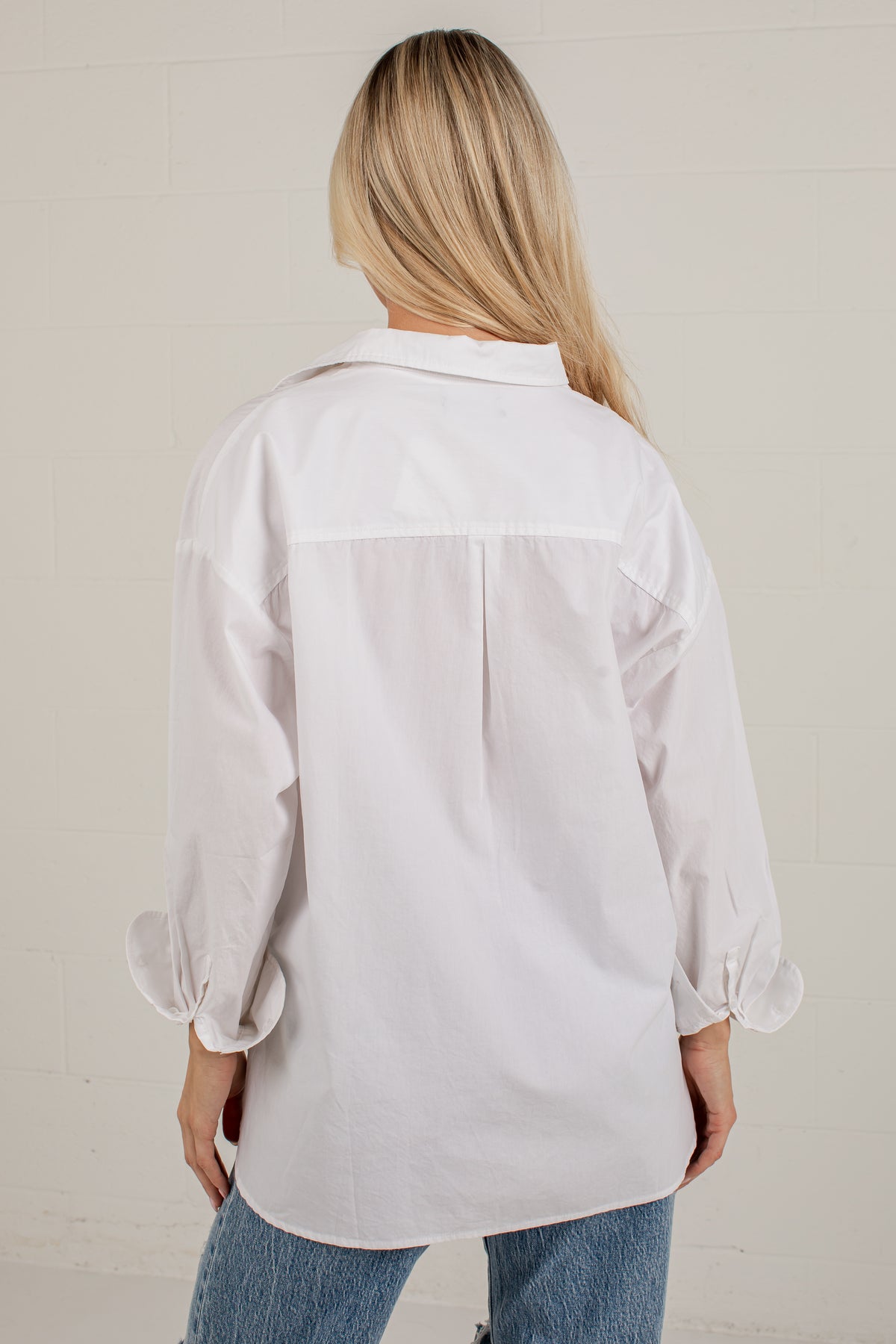 A woman viewed from behind wearing an oversized white button-up shirt with cuffed sleeves and blue jeans, standing against a light off-white brick wall.
