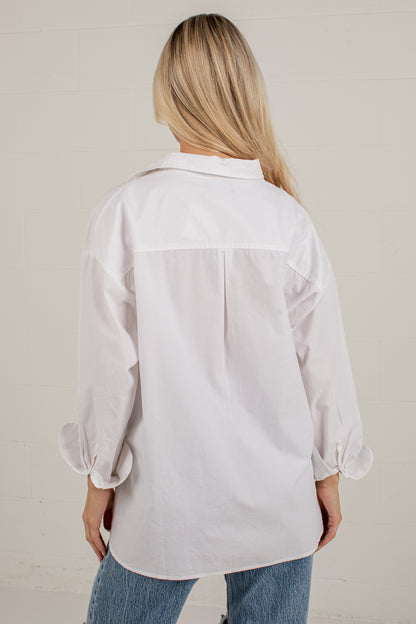 A woman viewed from behind wearing an oversized white button-up shirt with cuffed sleeves and blue jeans, standing against a light off-white brick wall.