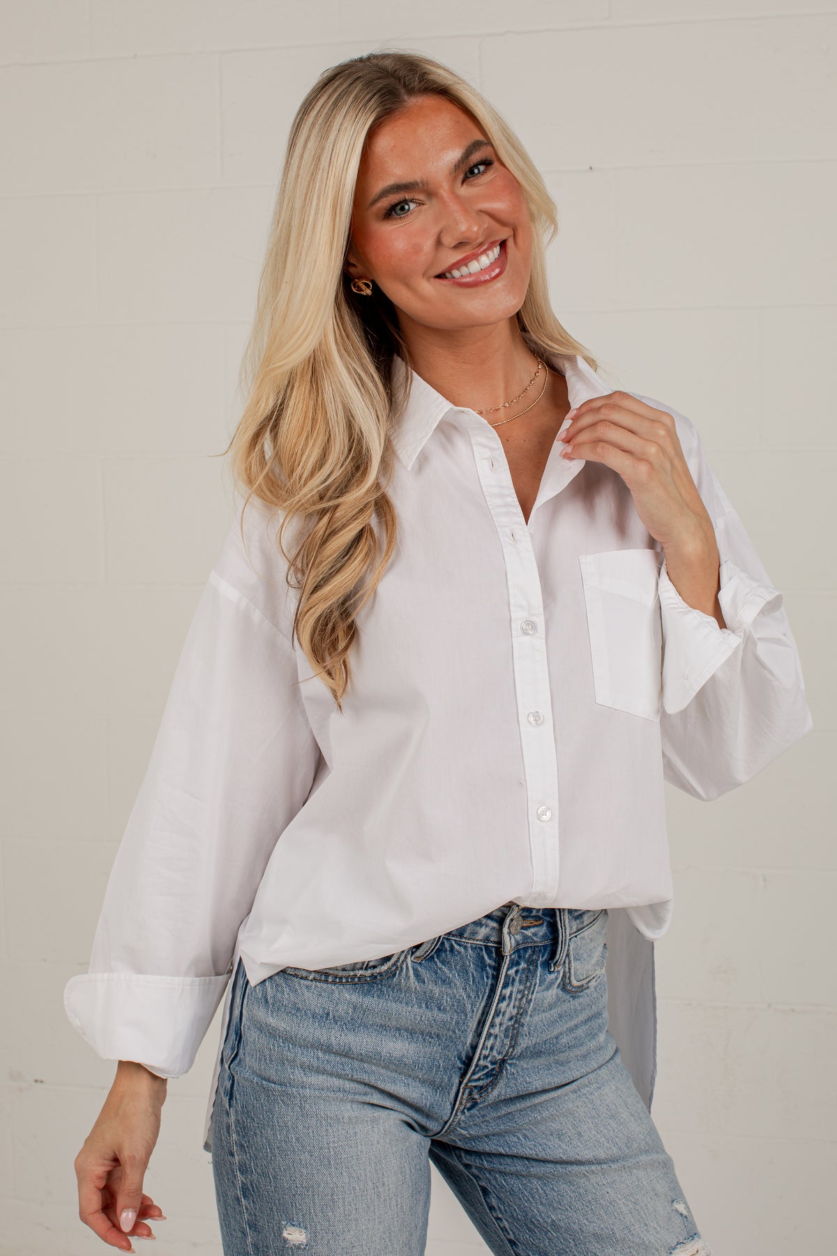 A smiling blonde woman wearing a white long-sleeve button-up shirt with rolled cuffs and light-wash blue jeans, standing against a light off-white brick wall.