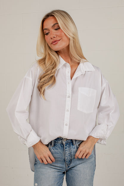 A woman wearing a white long-sleeve button-up shirt with a chest pocket and light blue jeans, standing against a light-colored brick wall.