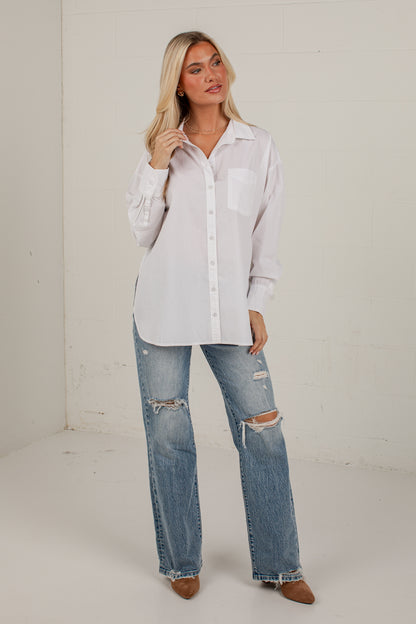 A woman modeling a white long-sleeve button-up shirt with a chest pocket, paired with distressed blue jeans and brown pointed-toe flats, standing against a white studio wall.