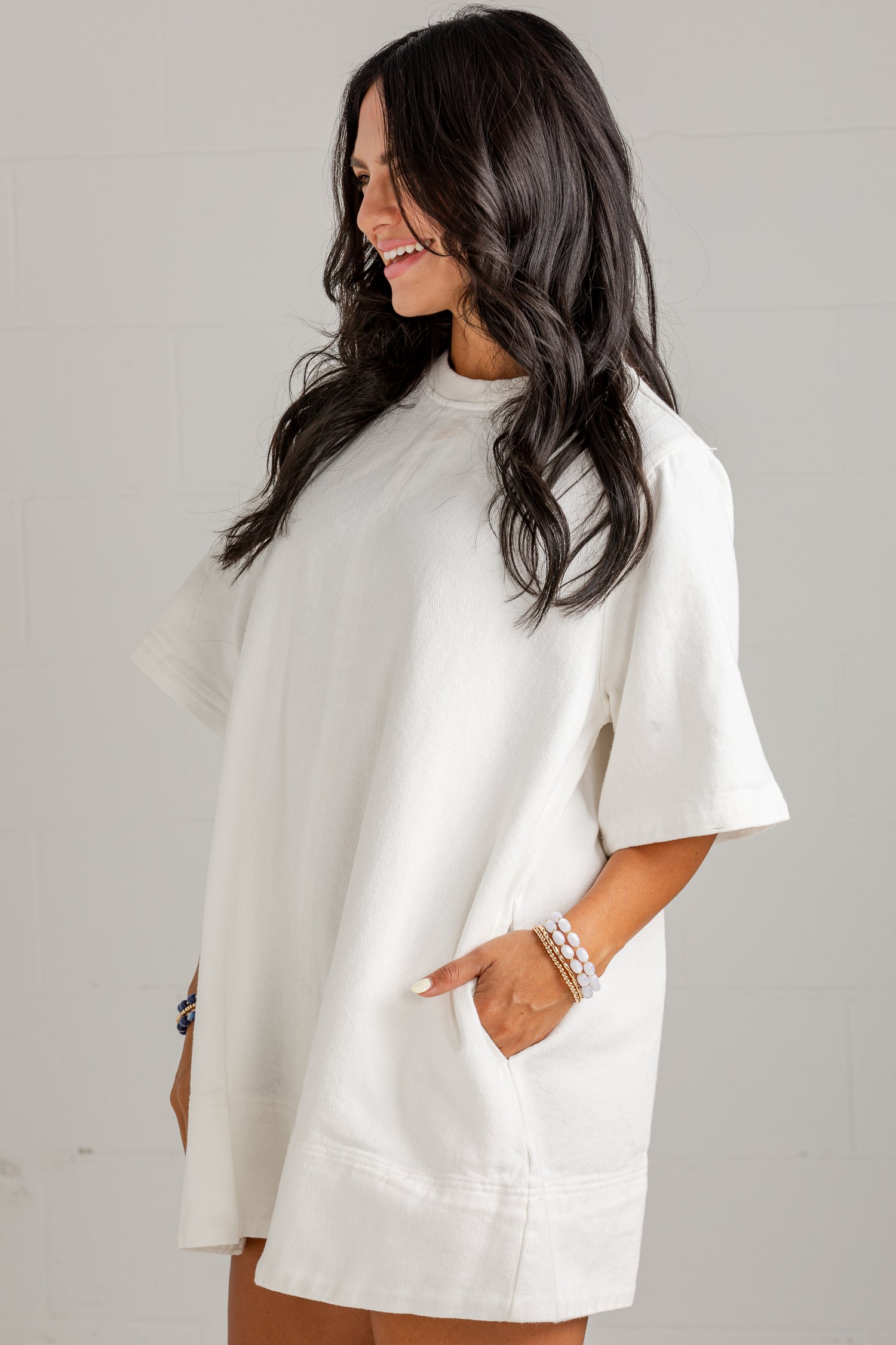 A woman wearing an oversized white short-sleeve tunic top with side pockets, standing against a light gray wall.