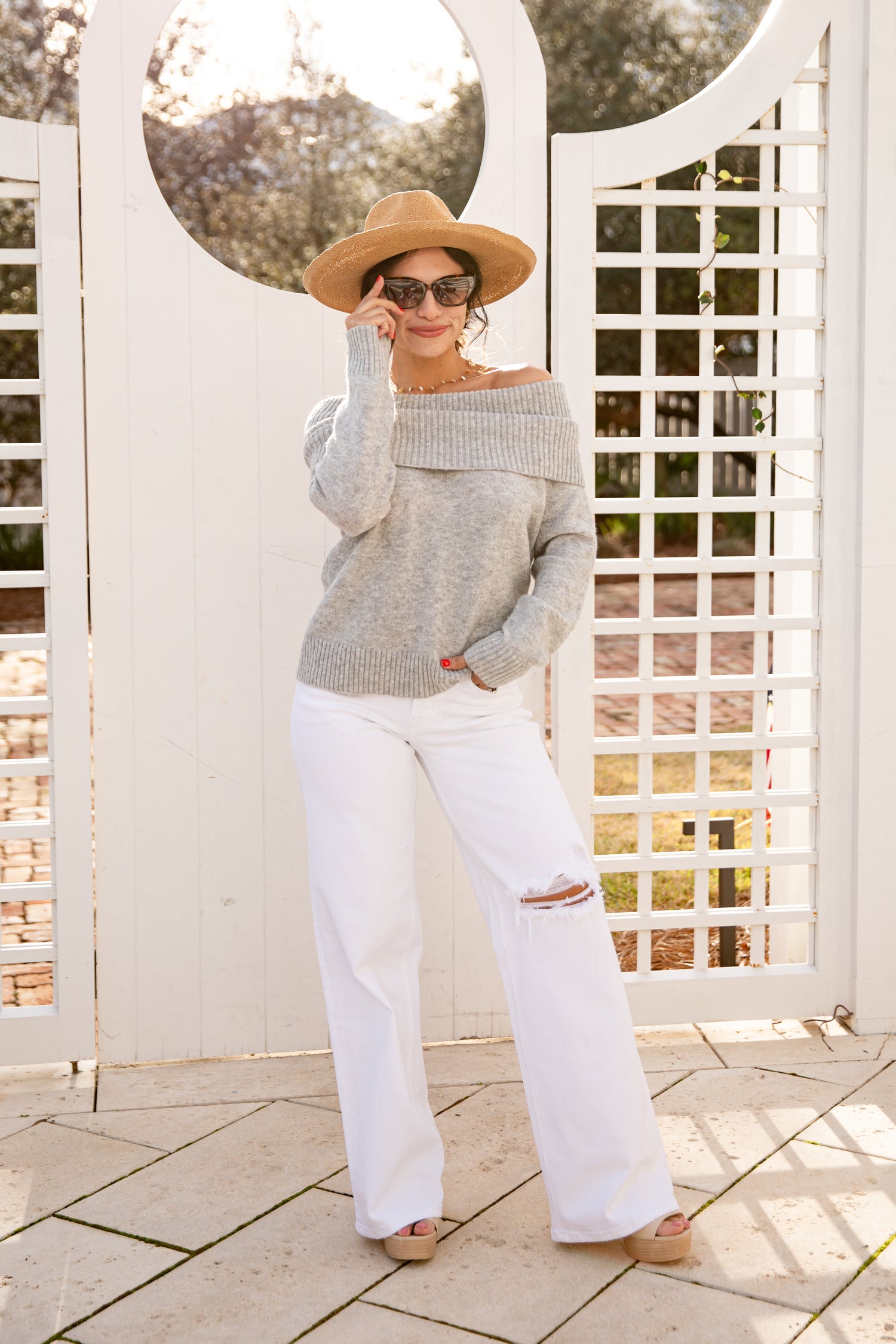 Woman wearing a gray sweater and white pants standing in front of a white lattice structure.