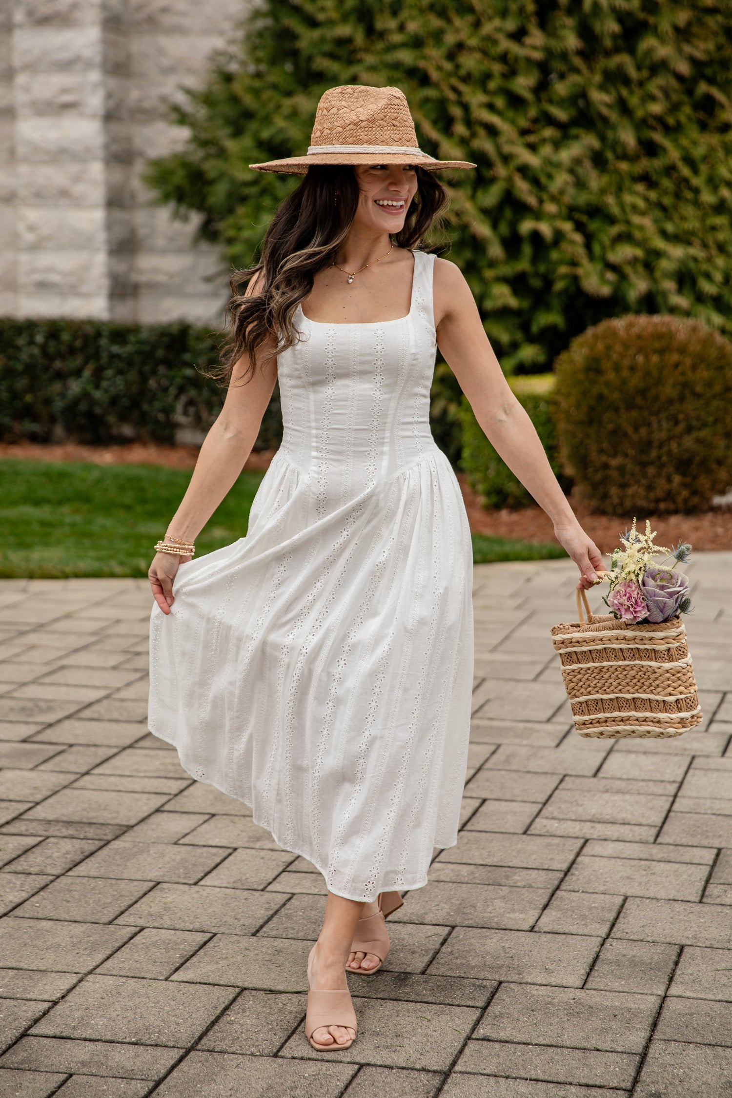 Woman in a white dress and straw hat holding a basket with flowers on a paved walkway.