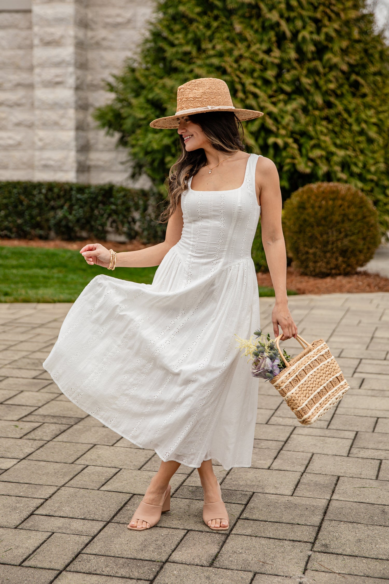 Woman in a white dress and straw hat holding a woven bag on a paved walkway.