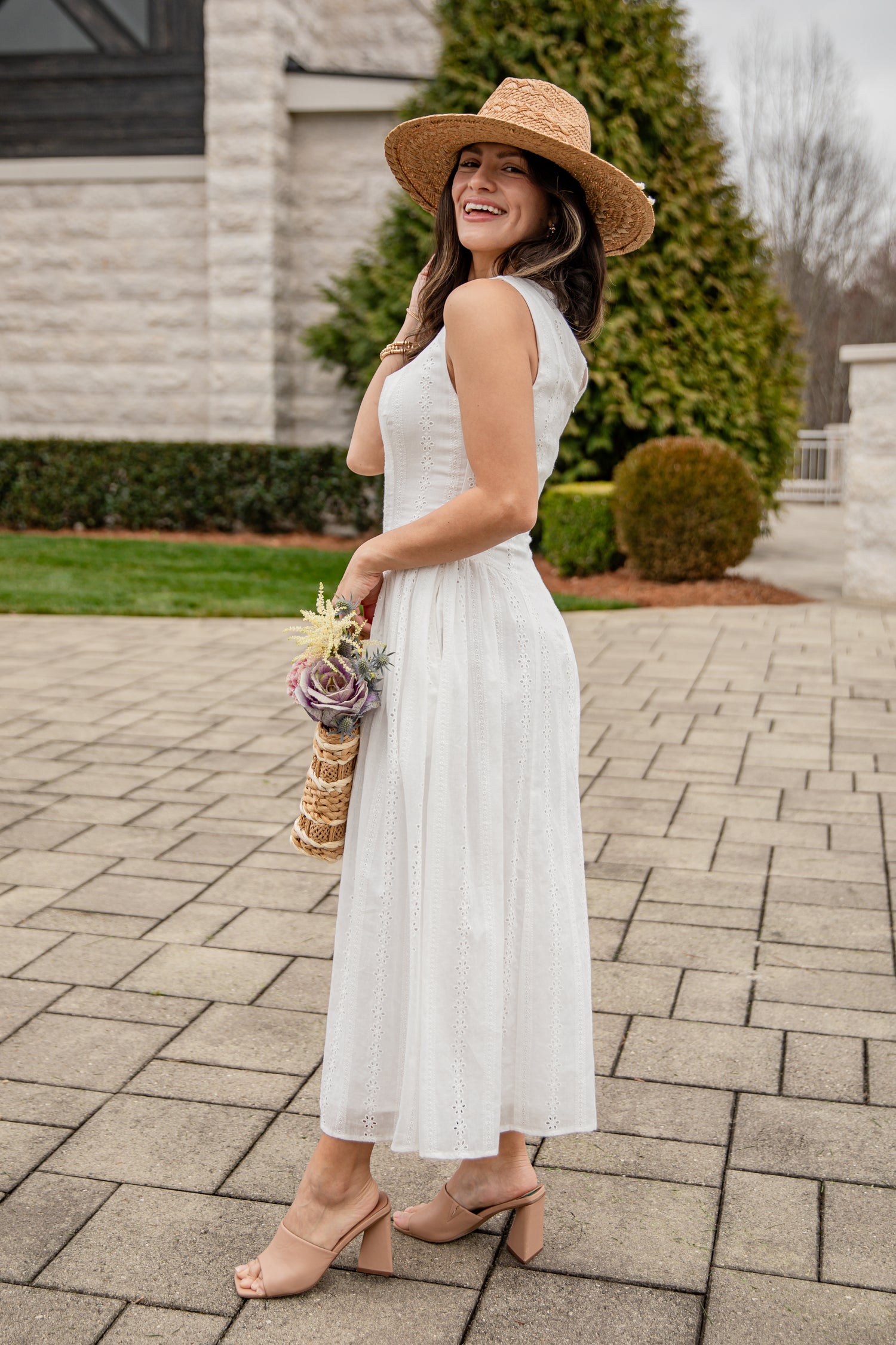 Woman in a white dress and straw hat standing on a paved walkway with greenery in the background.