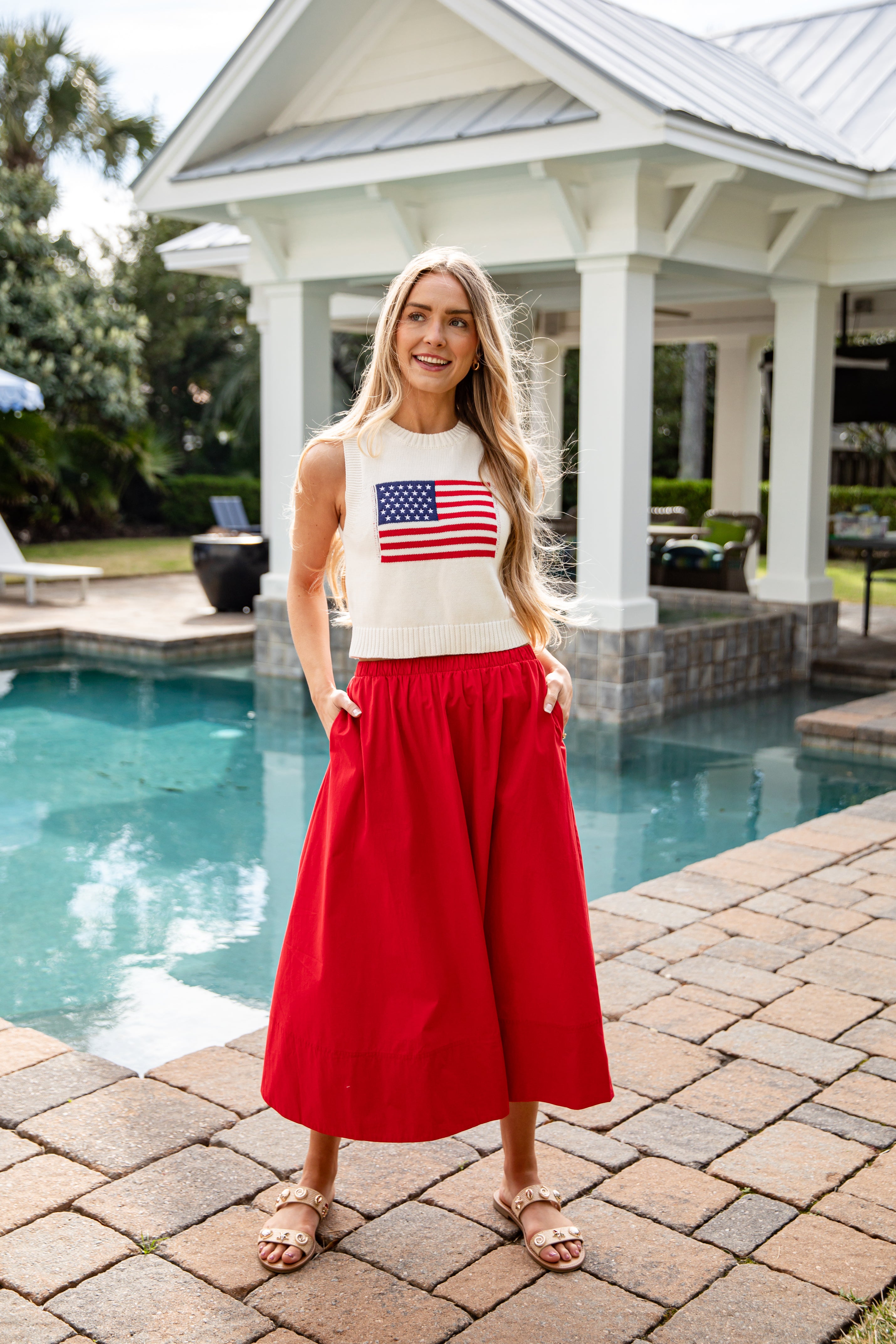 Woman in a white sleeveless top with an American flag design and red pants standing by a pool.