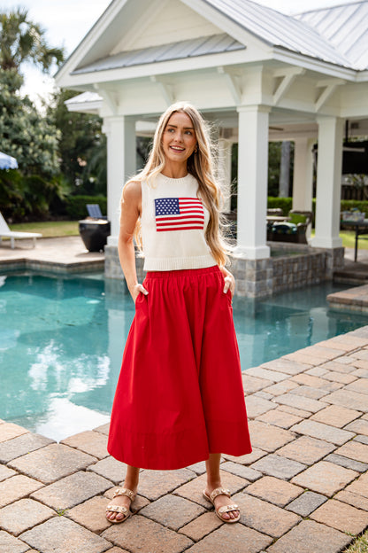 Woman in a white sleeveless top with an American flag design and red pants standing by a pool.