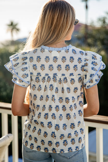 Woman wearing a floral patterned top with ruffled sleeves, standing on a balcony.