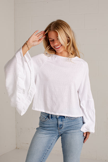 A woman wearing a white long-sleeve crop top with dramatic ruffled bell sleeves and light-wash blue jeans, standing against a white brick wall.