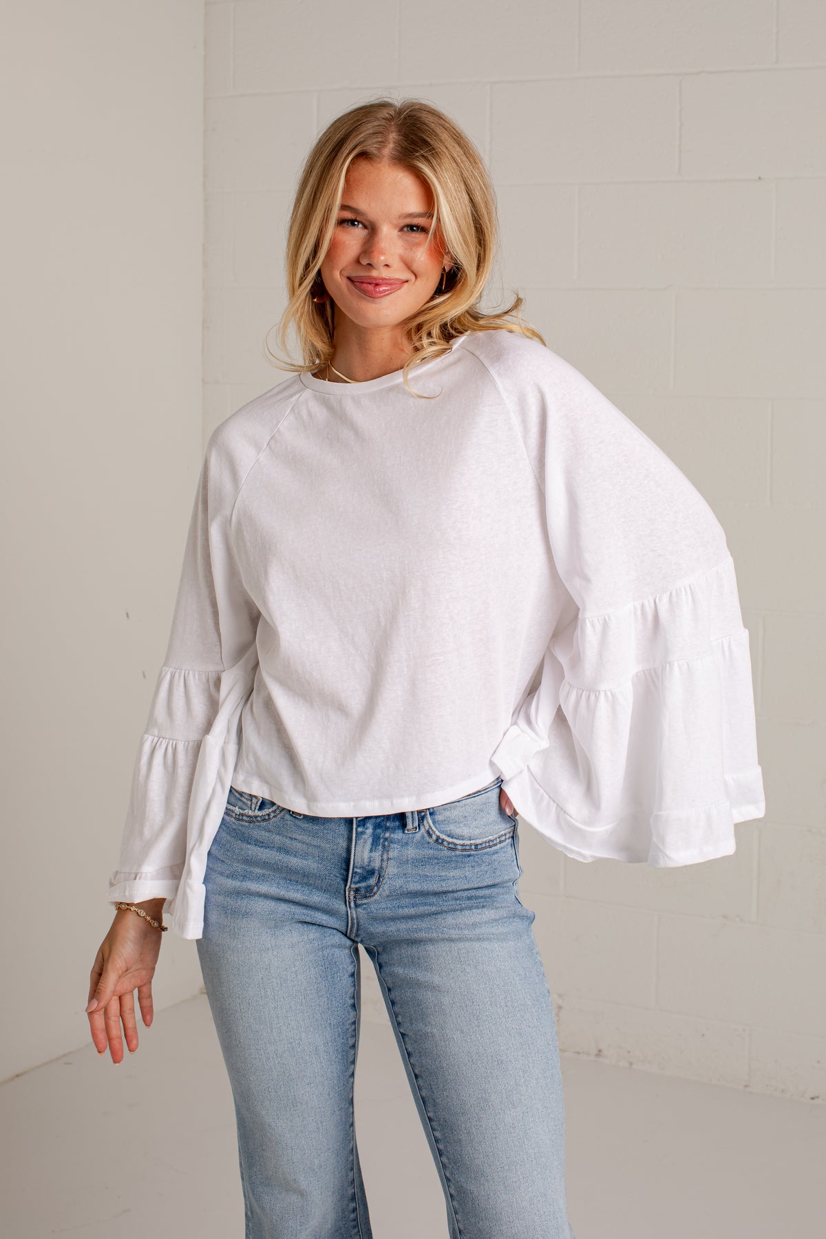 A woman wearing a white long-sleeve blouse with tiered ruffle sleeves and light blue jeans, standing against a white brick wall.