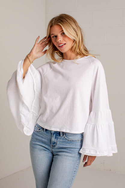 A woman wearing a white long-sleeve top with dramatic bell sleeves and light blue jeans, standing against a light gray wall.