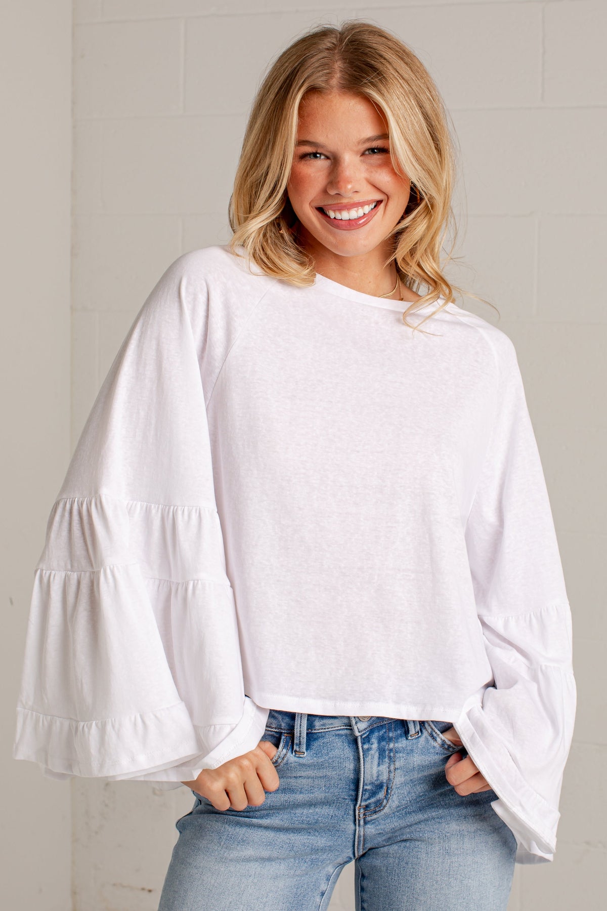 A smiling blonde woman wearing a white long-sleeve top with tiered ruffle sleeves and light blue jeans, standing against a light gray brick wall.