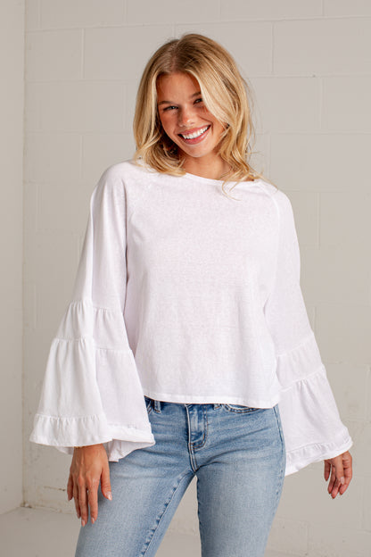 A smiling blonde woman wearing a white long-sleeve blouse with tiered ruffle bellows and light-wash blue jeans, standing against a light gray brick wall.