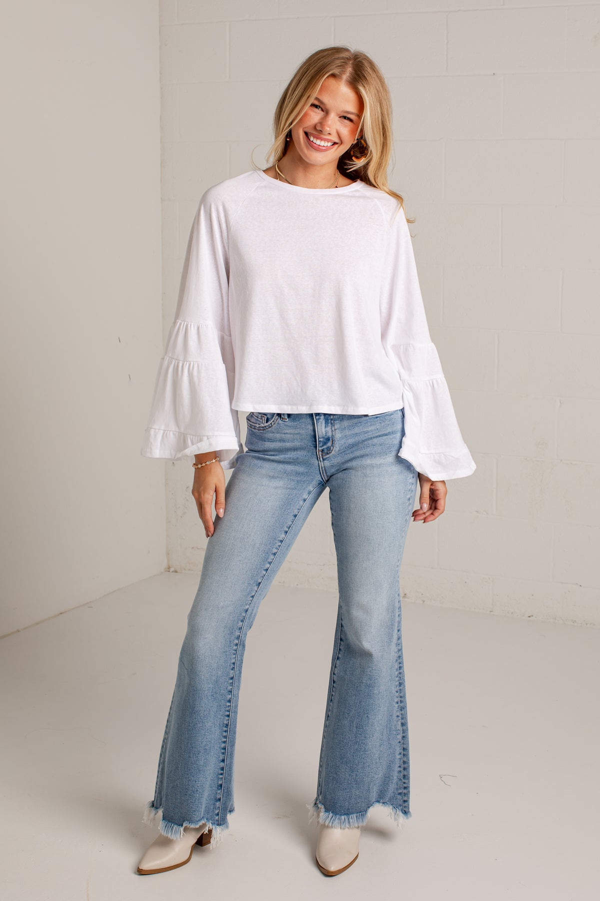 A woman modeling a white long-sleeve blouse with dramatic ruffled bell sleeves and light-wash blue flare jeans, standing against a plain off-white wall.
