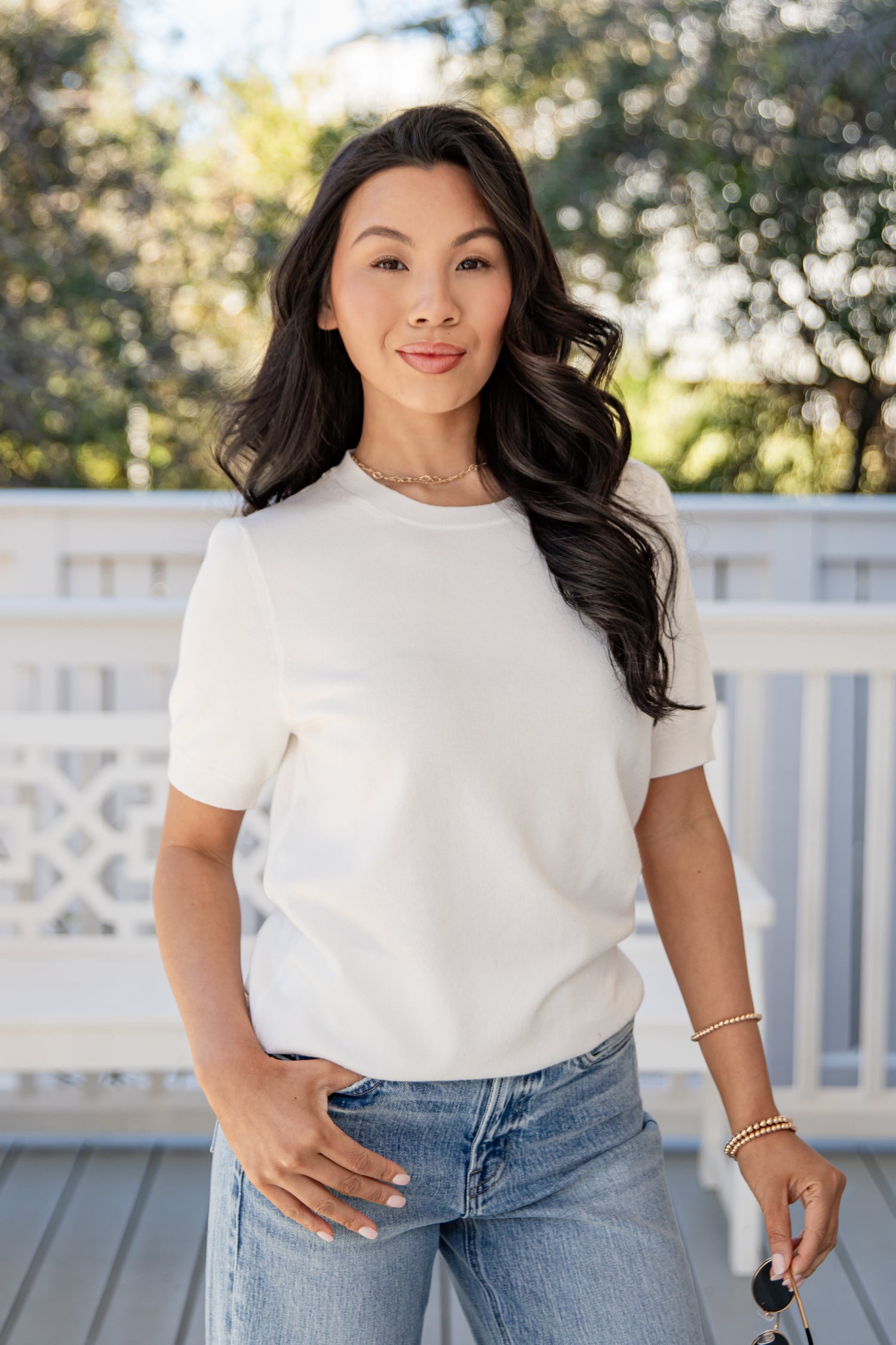 Woman wearing a white top and blue jeans standing on a wooden deck with greenery in the background