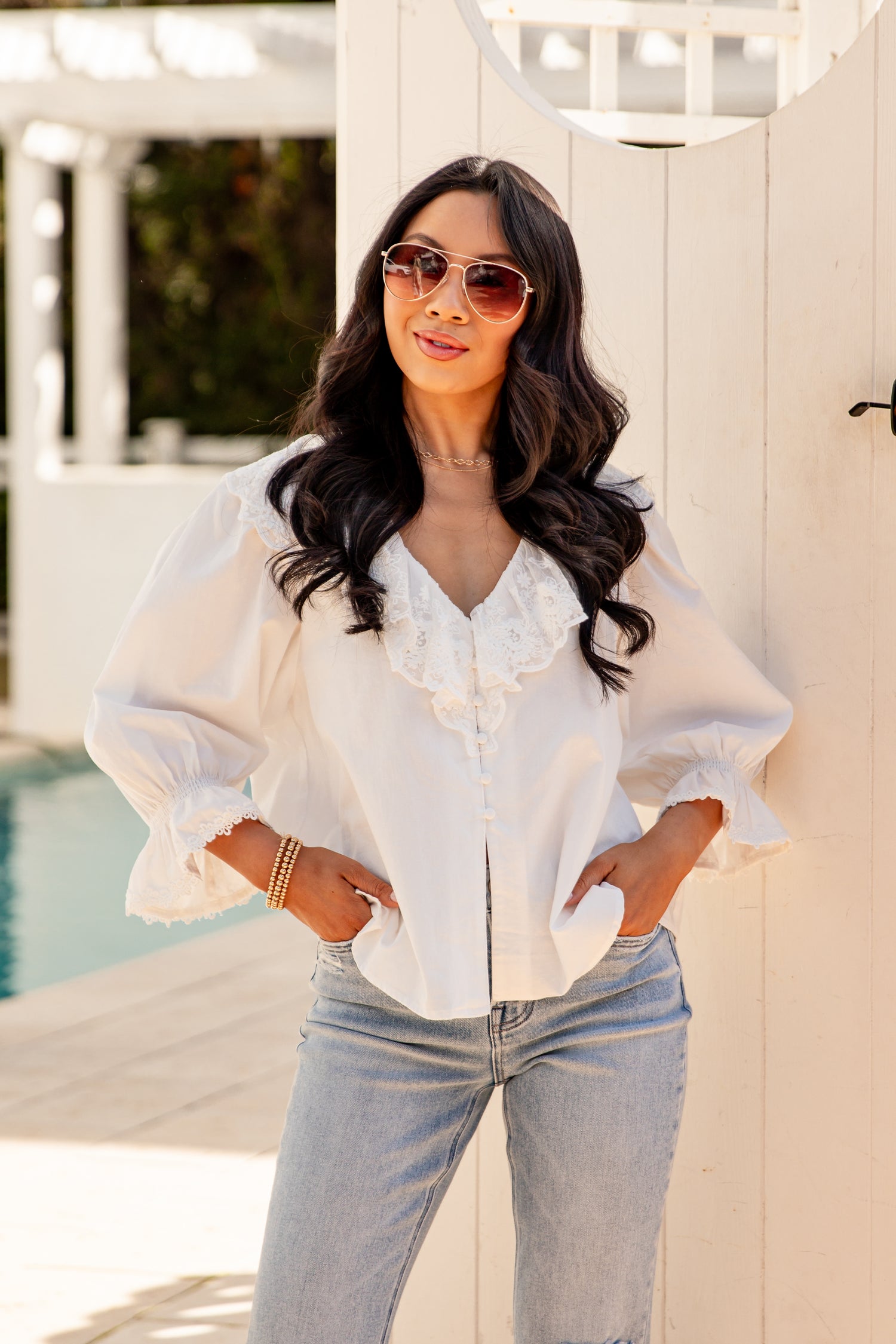Woman wearing a white blouse and sunglasses standing by a poolside.