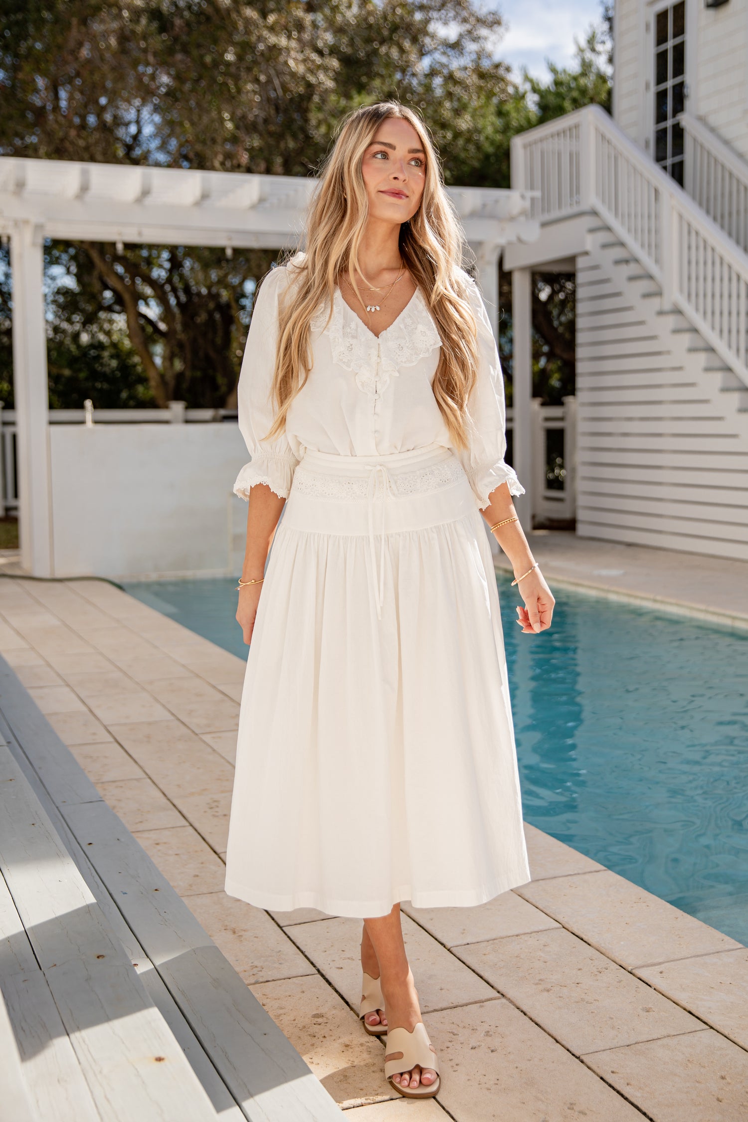 Woman in a white dress standing by a pool with trees and a house in the background