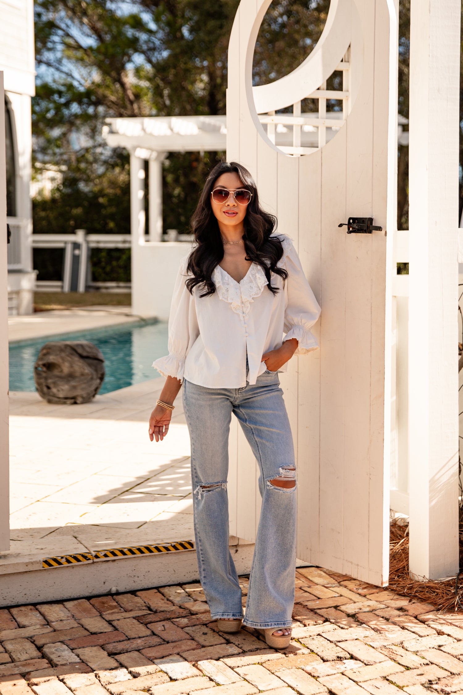 Woman in white blouse and jeans standing by a poolside with a white pergola in the background.