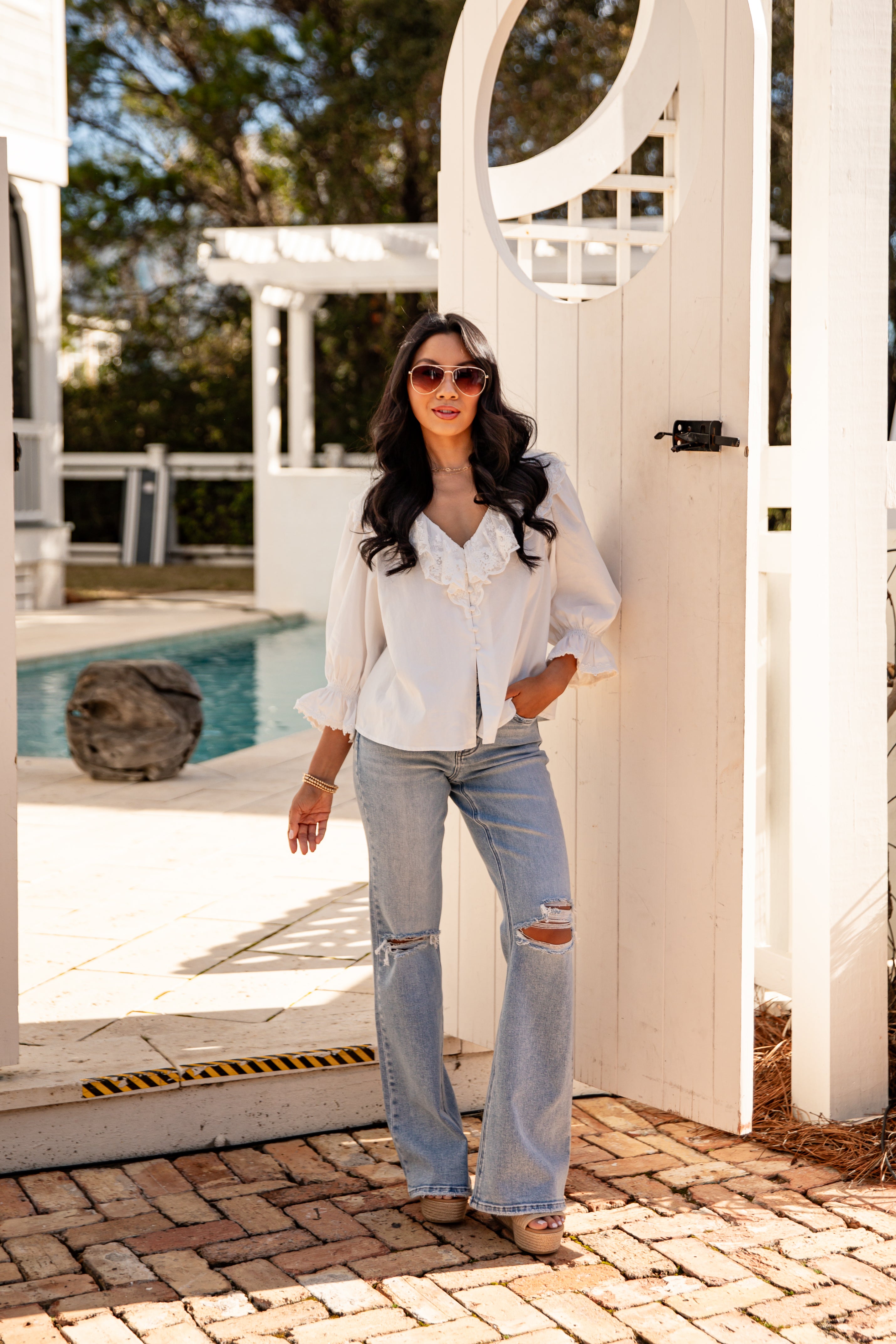 Woman in white blouse and jeans standing by a poolside with a white pergola in the background.
