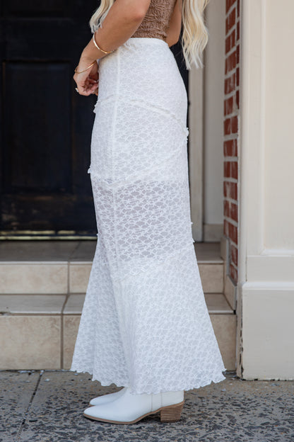 A woman wearing a long white lace maxi skirt with a textured floral pattern, paired with white ankle boots, standing on stone steps in front of a brick wall.