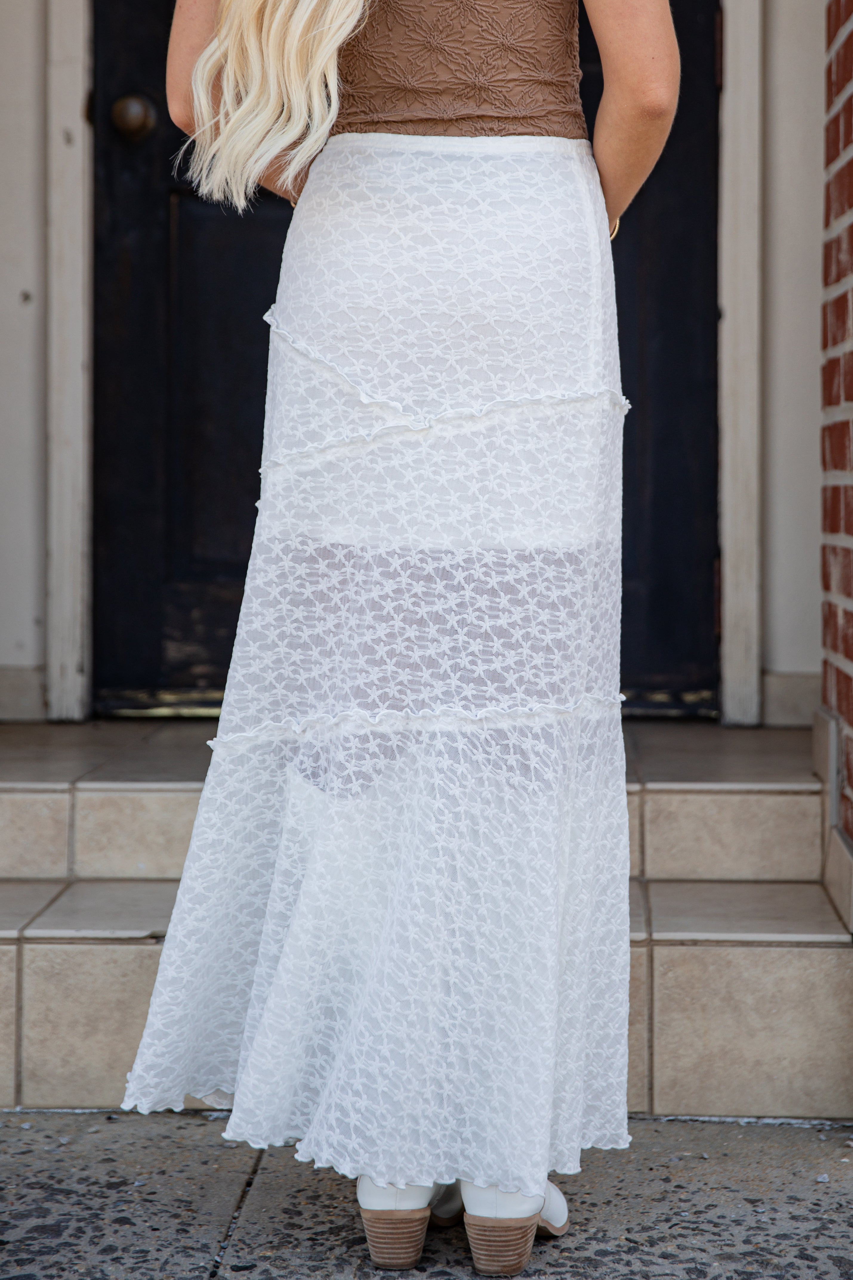 A woman wearing a long white lace maxi skirt with a tiered ruffle design, paired with a brown top and white boots, standing in front of a dark doorway.