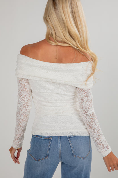 Person wearing a white off-shoulder top with lace sleeves and blue jeans on a plain background