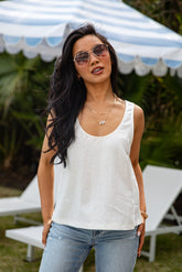Woman wearing a white sleeveless top and blue jeans with sunglasses, standing in front of lounge chairs and a striped canopy.