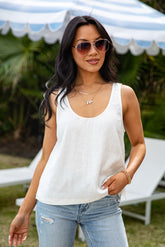 Woman wearing a white tank top and blue jeans outdoors with a striped canopy in the background.