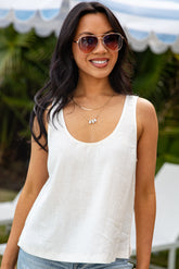 Woman wearing a white tank top and sunglasses outdoors with greenery in the background