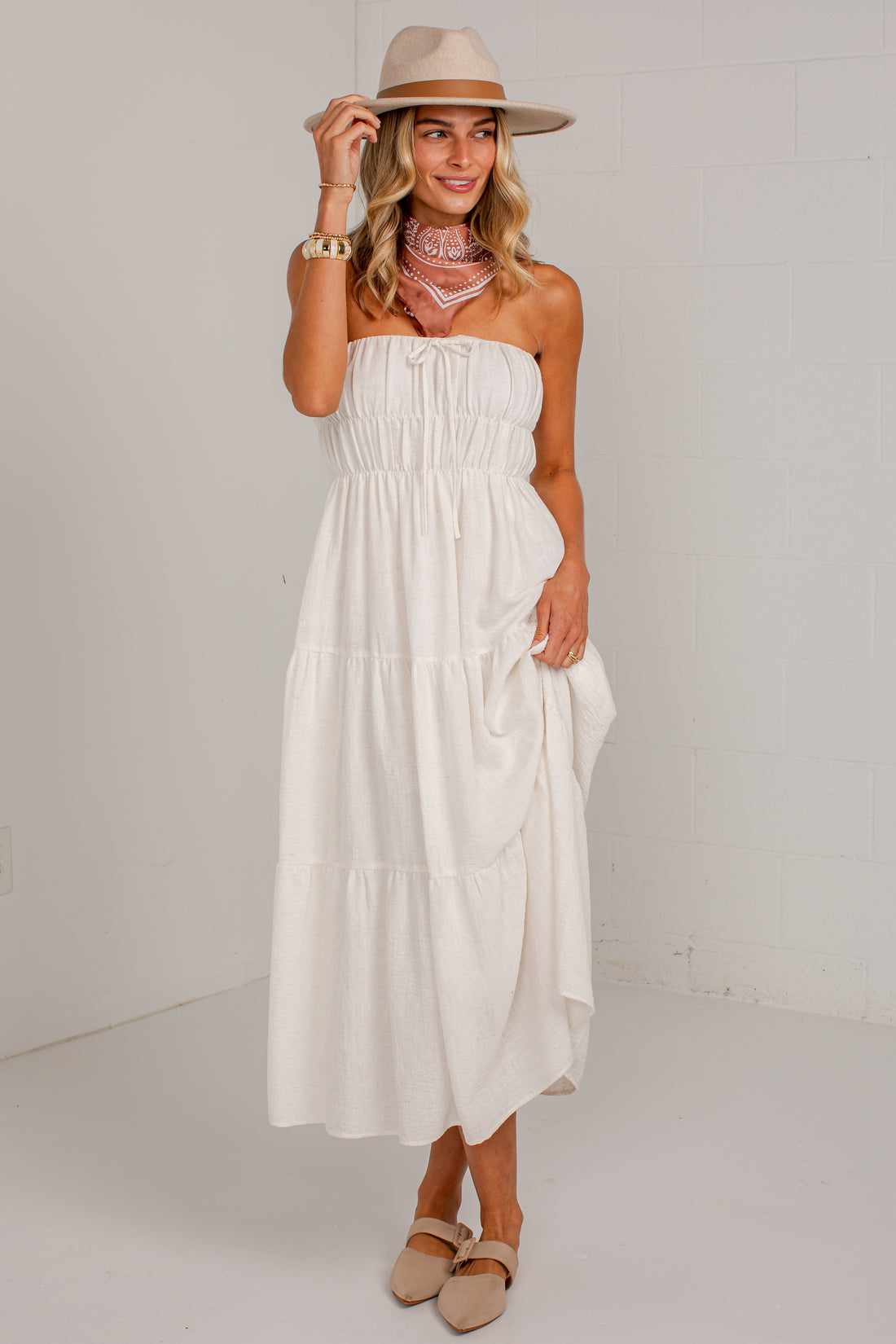 A woman modeling a strapless tiered midi dress in off-white, styled with a beige wide-brim hat, a pink patterned bandana, and beige flat shoes against a white studio wall.