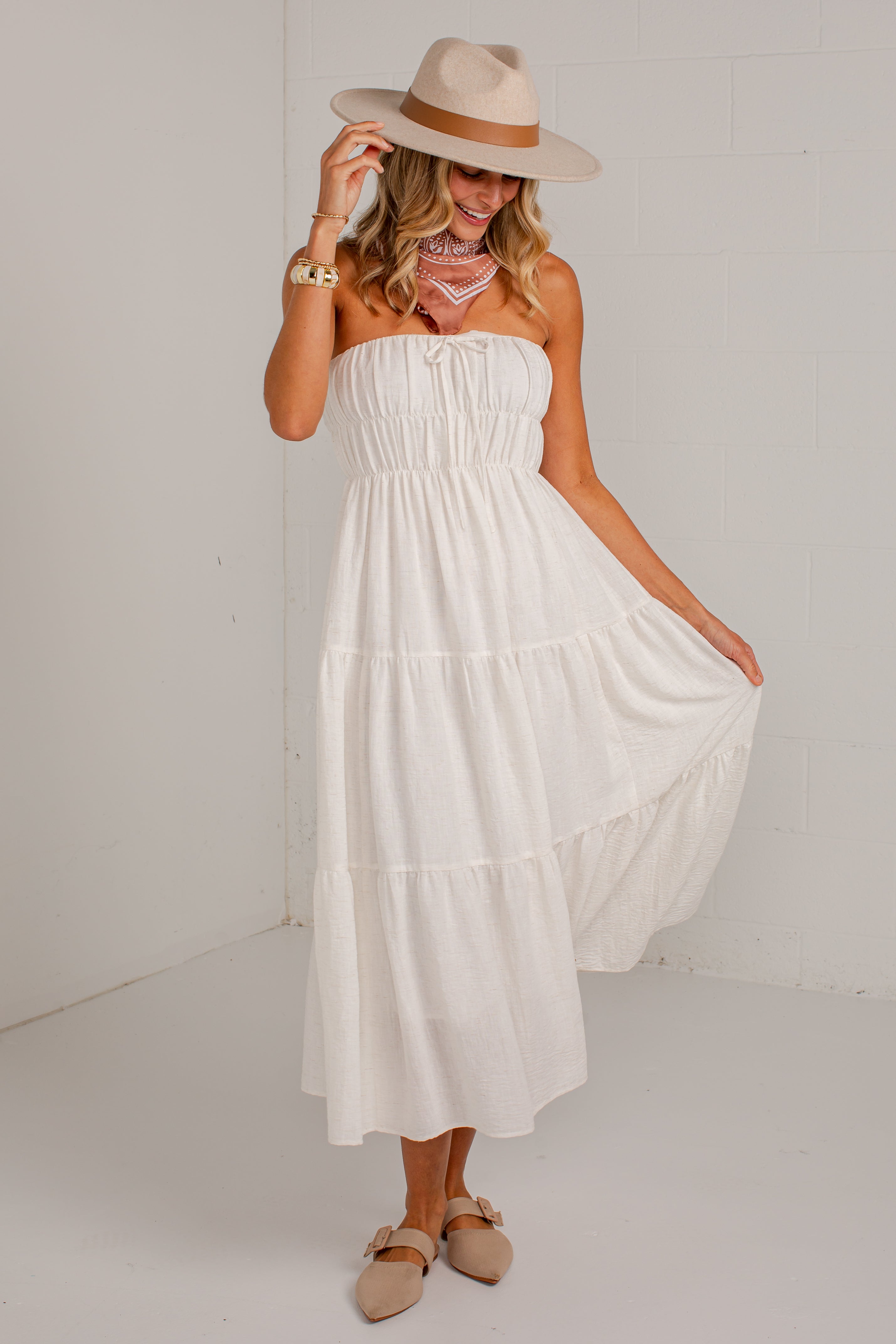 A woman wearing a strapless white tiered maxi dress, a beige wide-brim hat, and beige flat sandals, standing against a white wall.