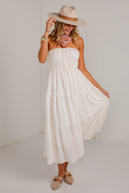 A woman wearing a strapless white tiered maxi dress, a beige wide-brim hat, and beige flat sandals, standing against a white wall.