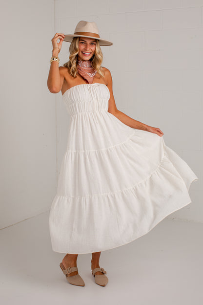 A woman modeling a white strapless tiered maxi dress with a smocked bodice, paired with a beige wide-brim hat, patterned scarf, and beige flat shoes against a white studio background.