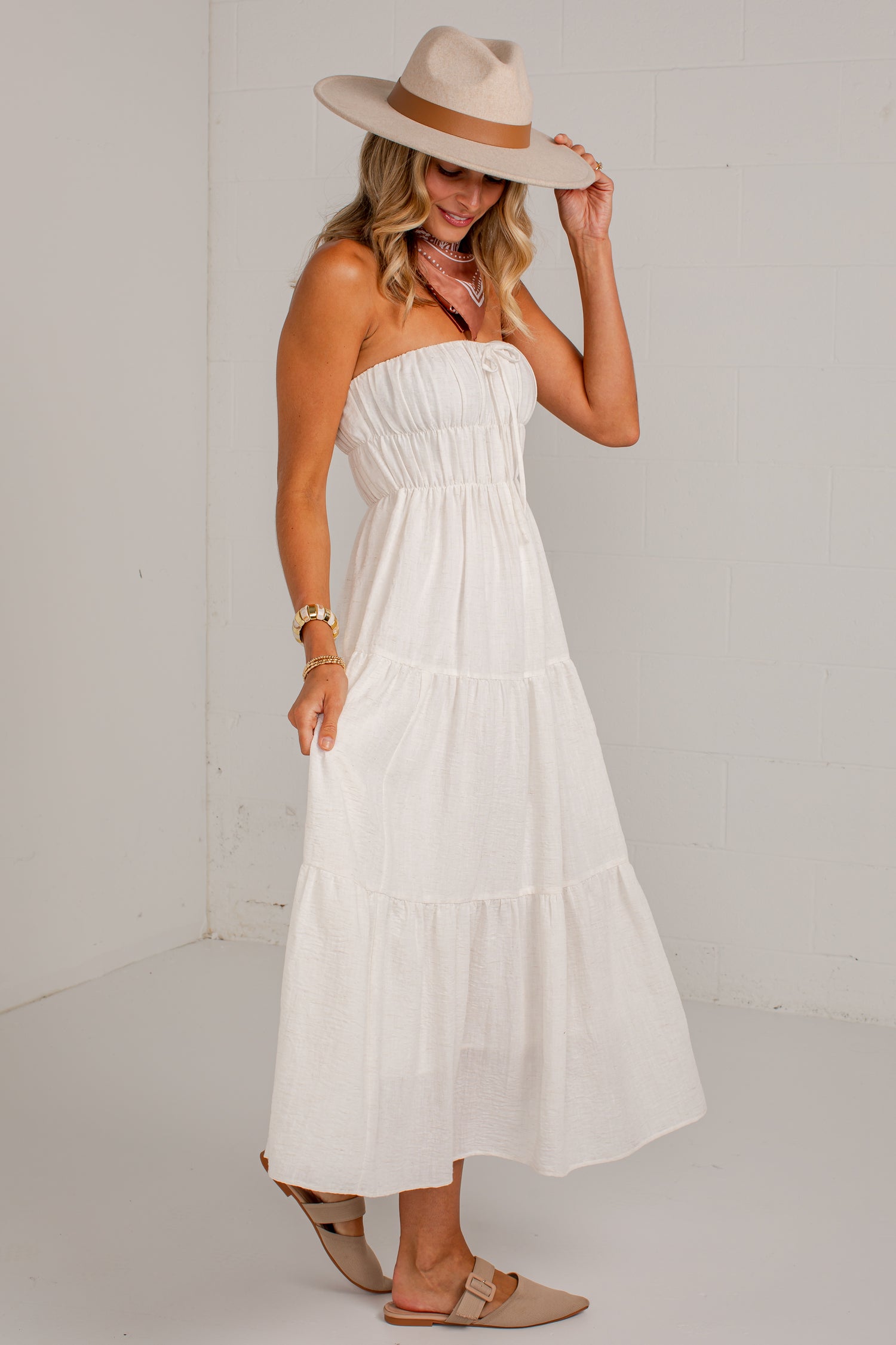 A woman wearing a strapless white tiered maxi dress, a beige wide-brim hat, and beige flat sandals, standing against a white wall.