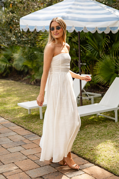 Woman in a strapless white dress standing under a striped umbrella in a garden setting.
