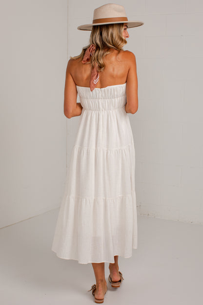 A woman wearing a strapless white tiered maxi dress, a beige wide-brim hat, and beige flat sandals, standing against a white wall.