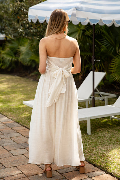 Woman in a white strapless dress with a bow standing under a striped umbrella in a garden setting.