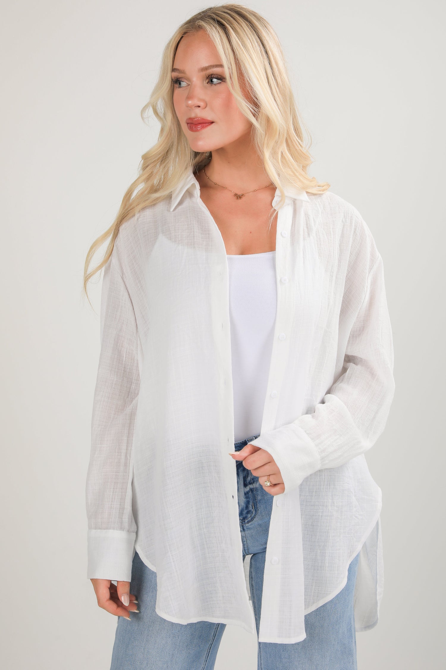 Woman wearing a white sheer blouse over a white top with blue jeans on a light gray background