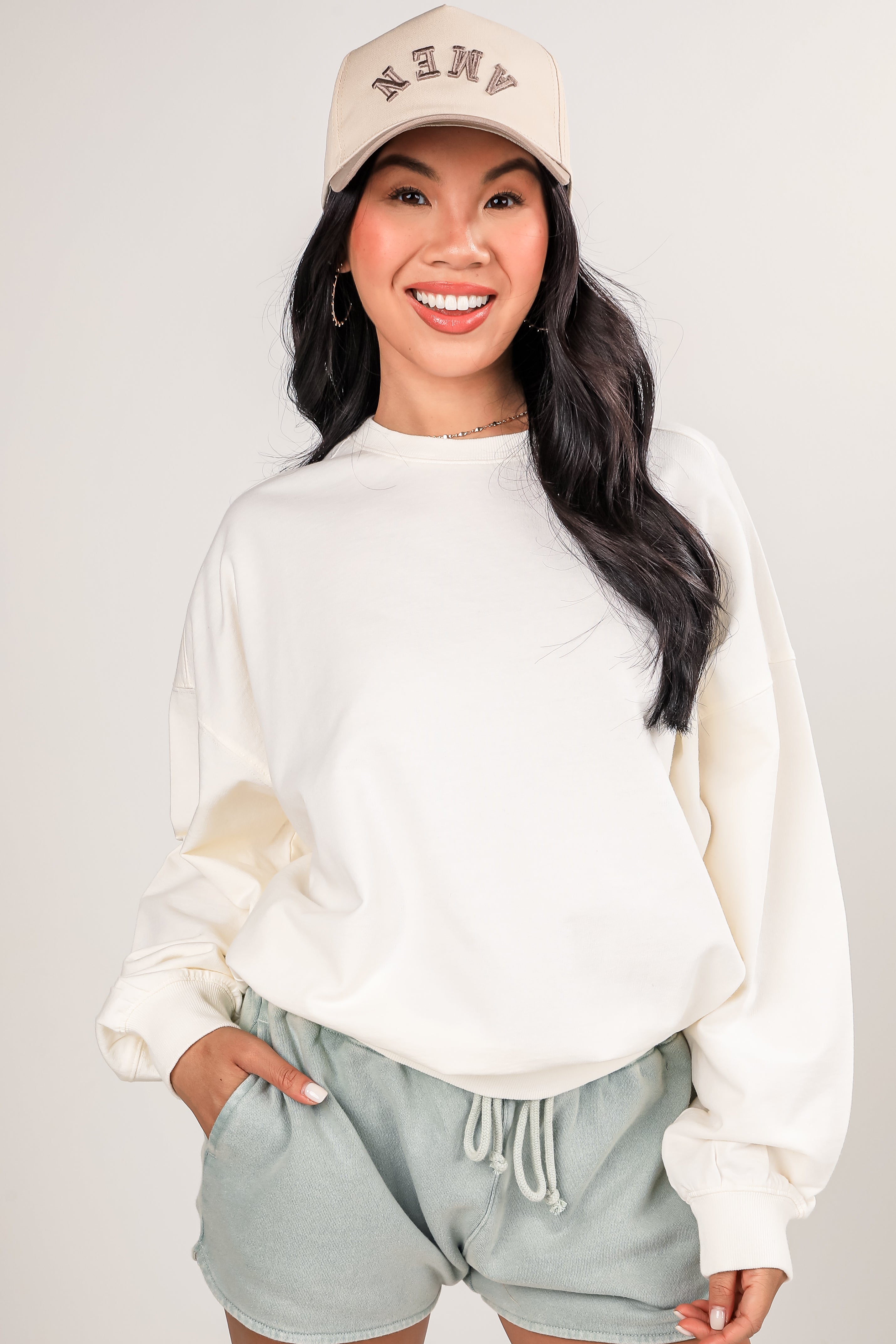 Woman wearing a beige cap and white sweatshirt with light gray shorts on a plain background