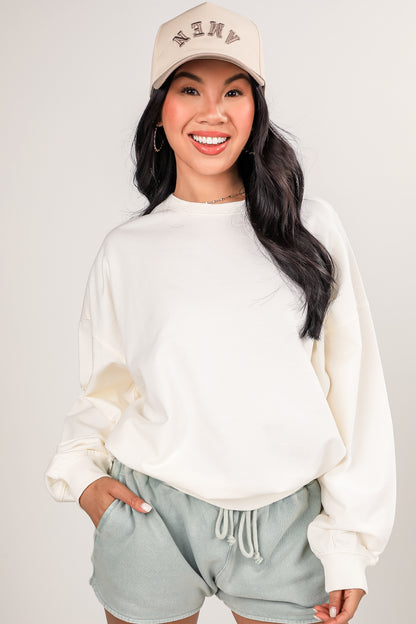 Woman wearing a beige cap and white sweatshirt with light gray shorts on a plain background