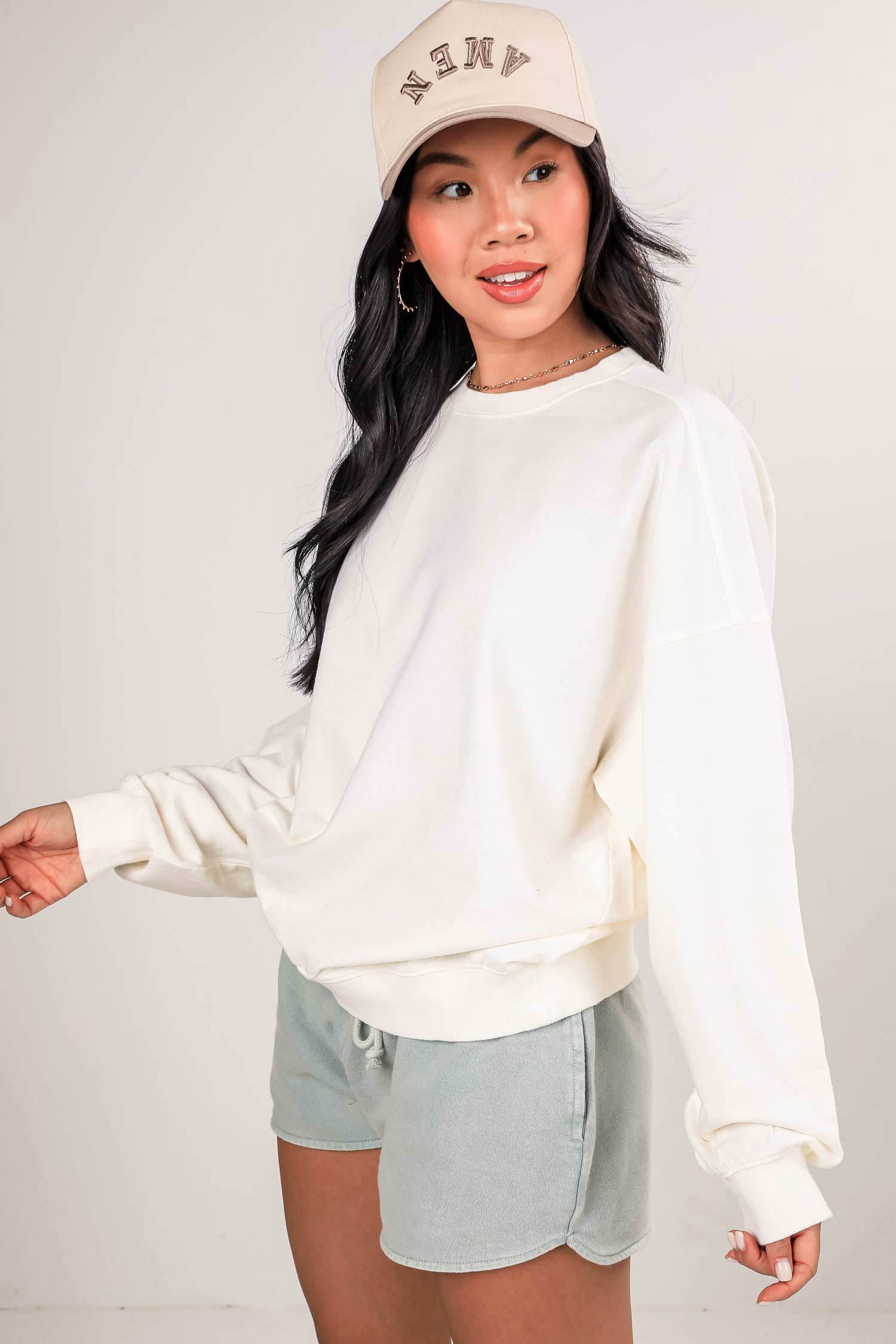 Woman wearing a white sweatshirt and light shorts with a beige cap on a plain background