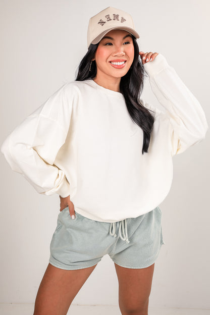 Woman wearing a white sweatshirt and light green shorts with a beige cap on a plain background