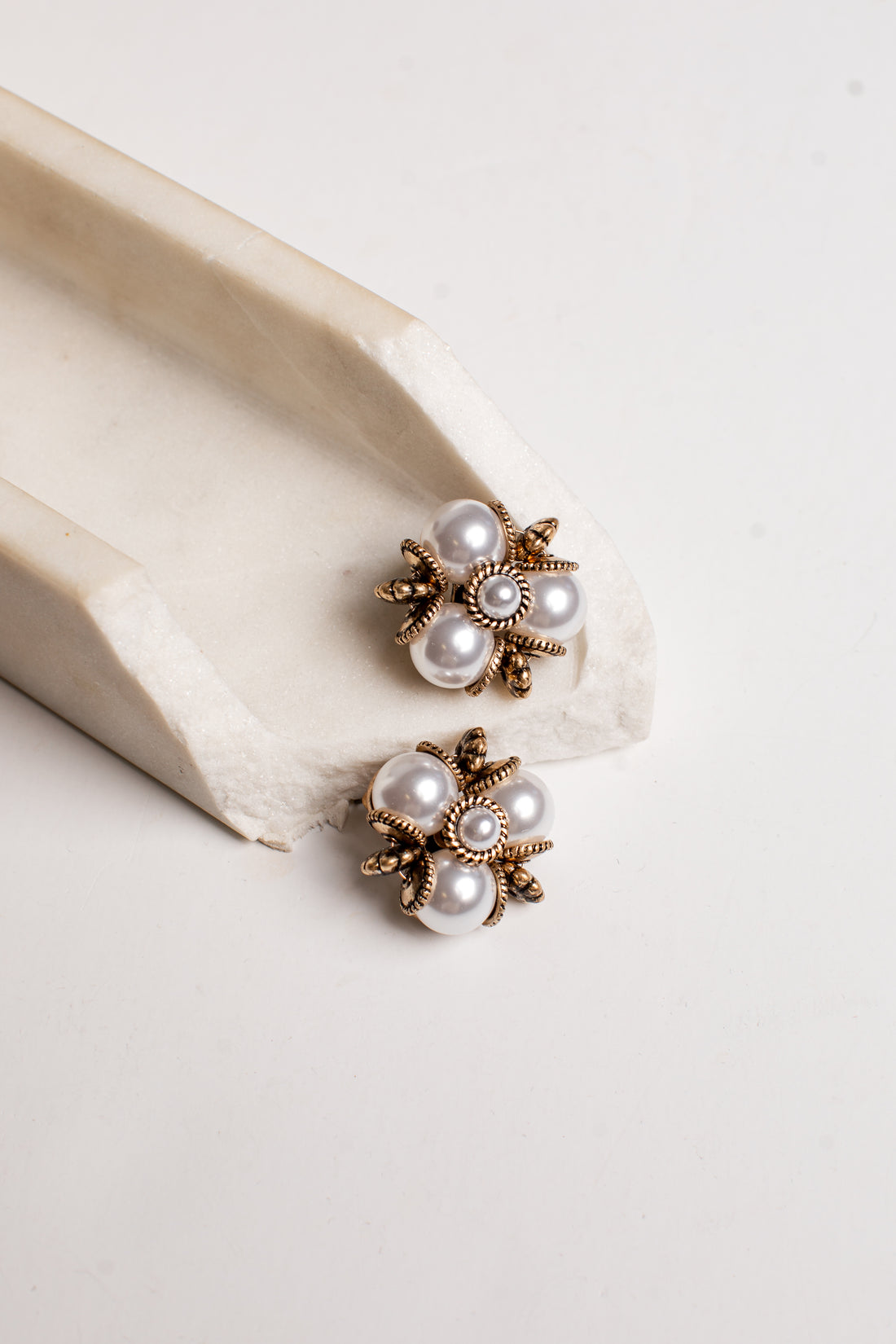 A pair of gold-toned flower-shaped earrings with white faux pearls, displayed on a cream-colored ceramic stand against a light off-white background.