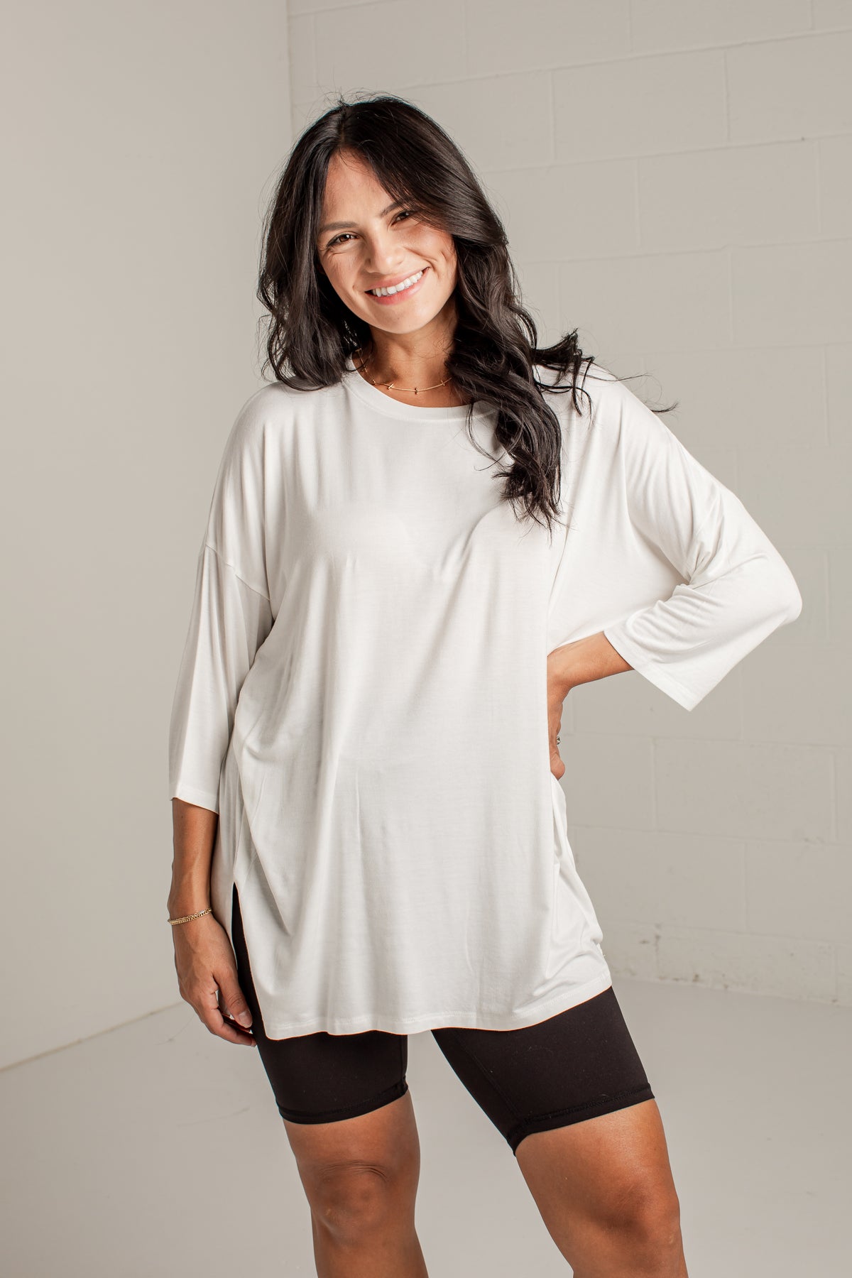 A woman modeling a white oversized long-sleeve tunic top with black fitted shorts against a light gray studio wall.