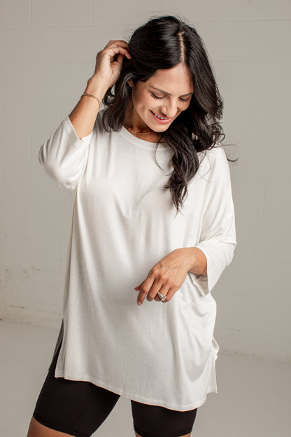 A woman wearing a loose-fitting white long-sleeve tunic top over black fitted shorts, standing against a light neutral wall.