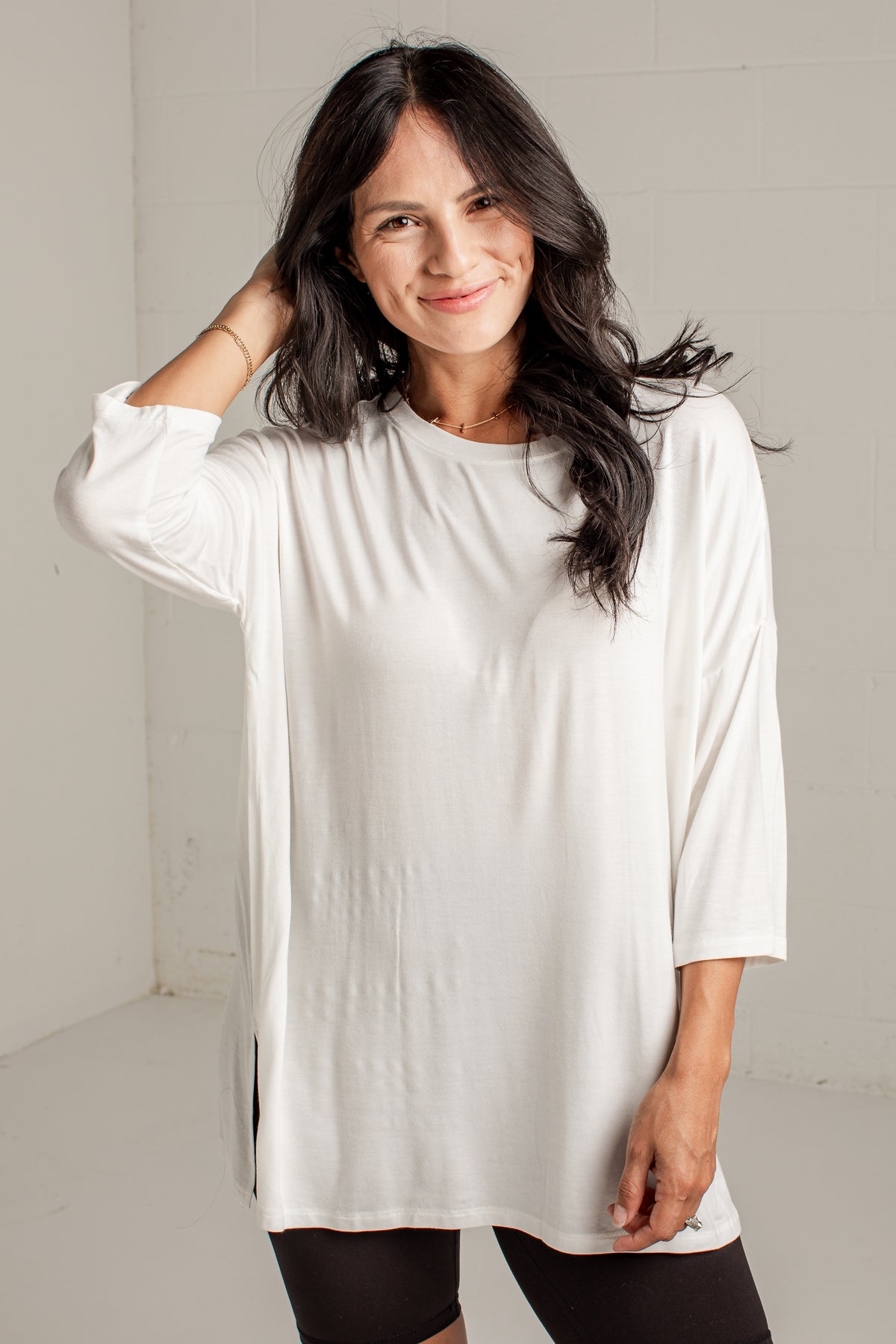 A woman with long dark hair wearing a loose-fitting white long-sleeve tunic top and black leggings, standing against a light gray wall.