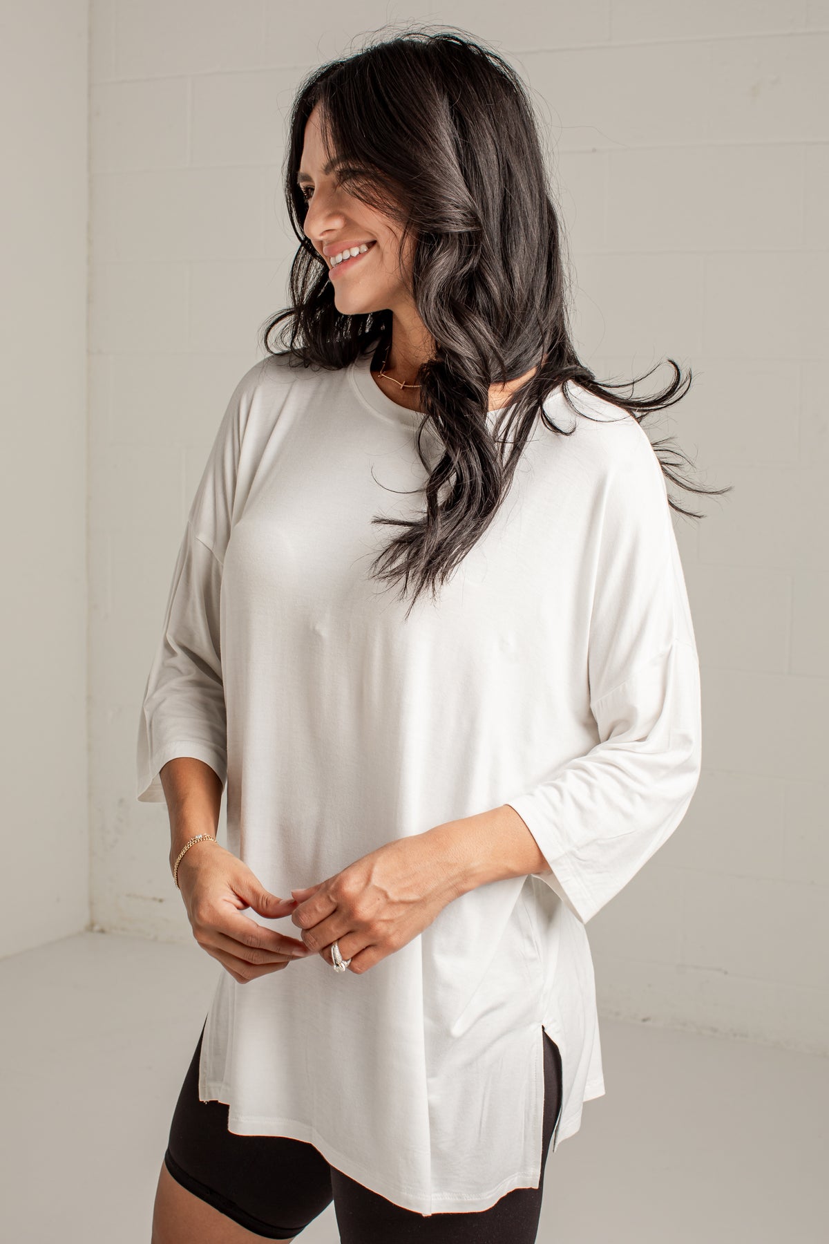 A woman wearing a loose white three-quarter sleeve top with side slits, paired with black shorts, standing against a white brick wall.