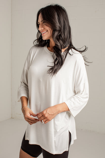 A woman wearing a loose white three-quarter sleeve top with side slits, paired with black shorts, standing against a white brick wall.